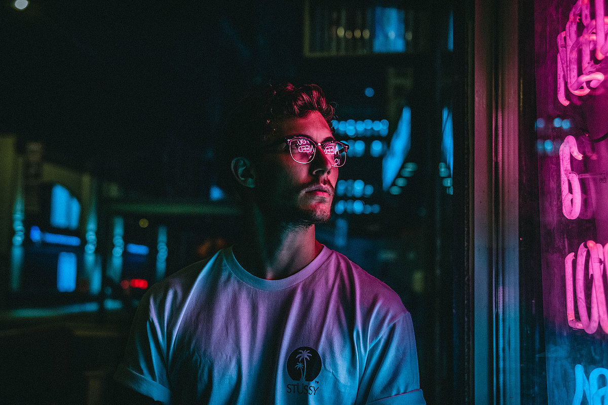 Junger Mann schaut mit Brille in ein Fenster und wird in NEON Lichtern angestrahlt